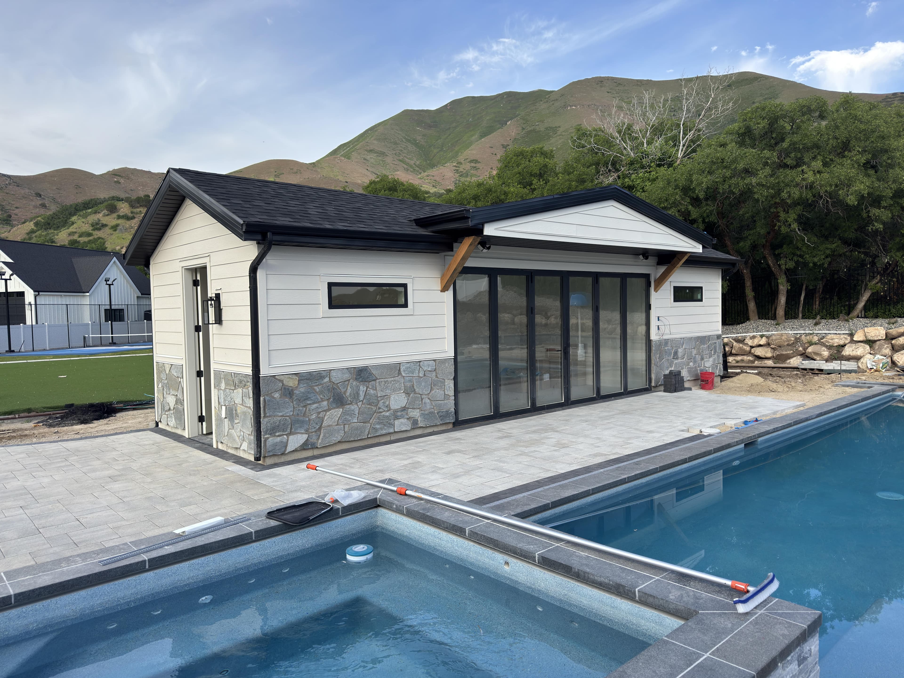 Modern pool house with large glass doors, stone accents, and a scenic mountain backdrop.