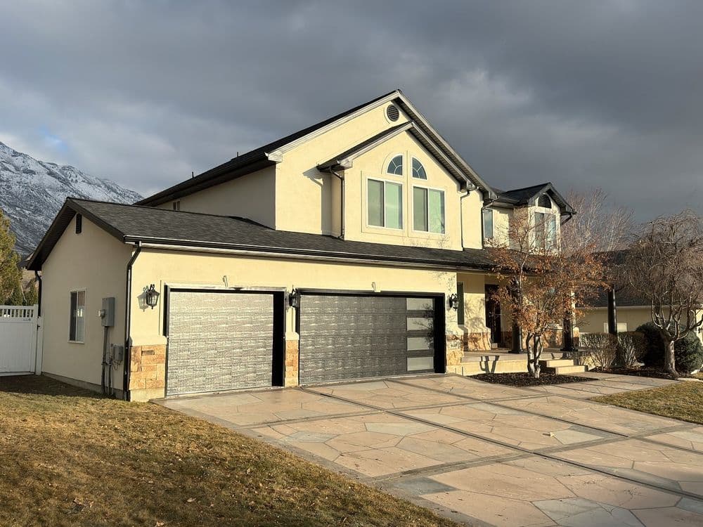 Two-story family home with double garage, stone pathway, and mountain backdrop under cloudy sky.