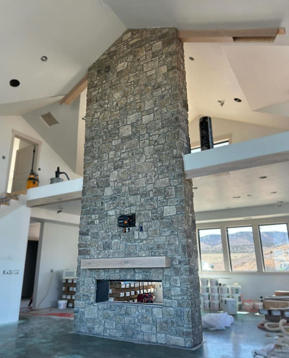 Stone fireplace with wooden mantel in modern home interior with large windows.
