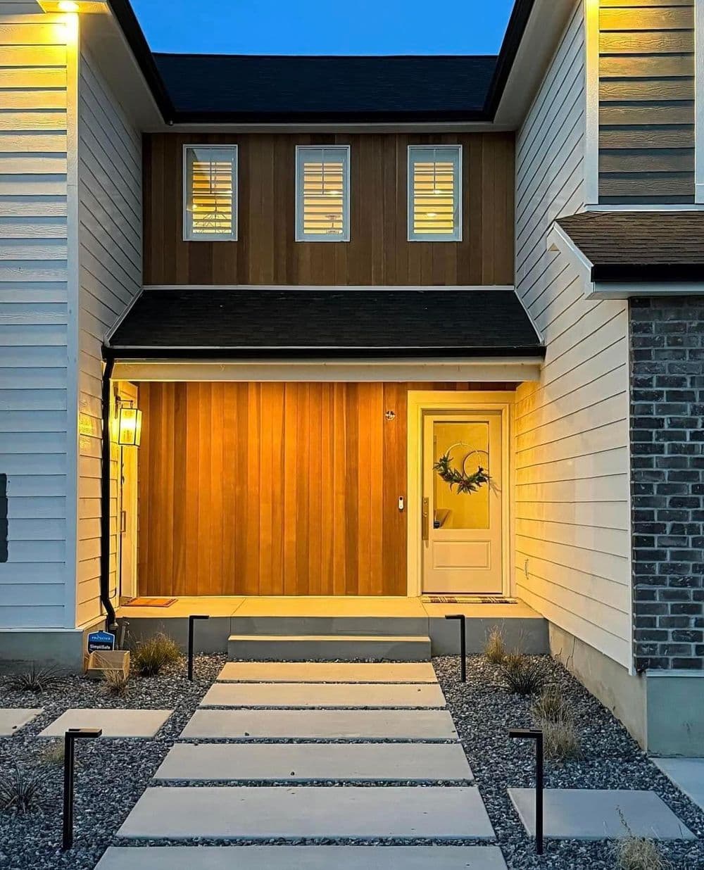 Modern home entrance with warm lighting, wooden accents, and stylish pathway at dusk.