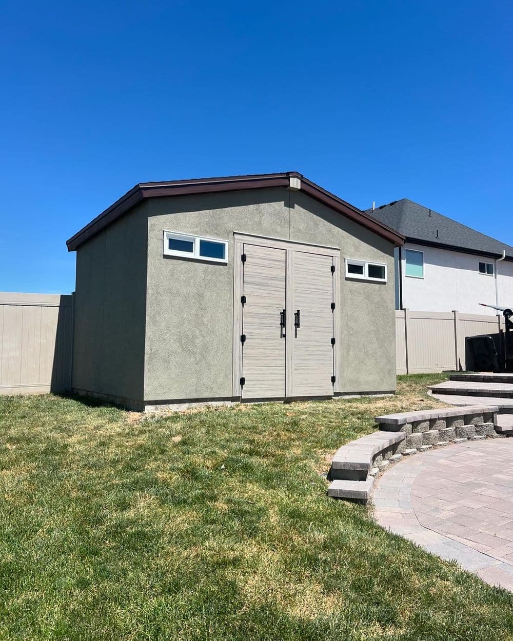 Modern outdoor storage shed with double doors and windows on a sunny day. Green grass and patio stones.