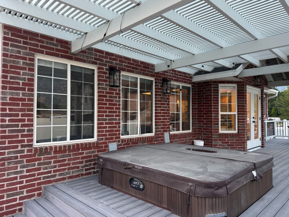 Hot tub on a wooden deck beneath a slatted awning, surrounded by brick walls and windows.