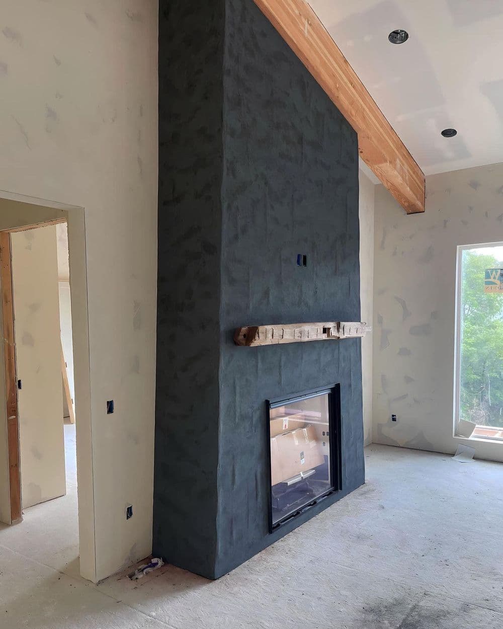 Modern living room with dark textured fireplace, wooden mantel, and unfinished drywall.