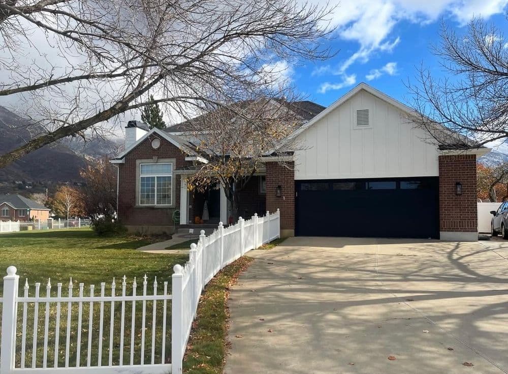 Modern brick home with a black garage door, white fence, and mountains in the background.